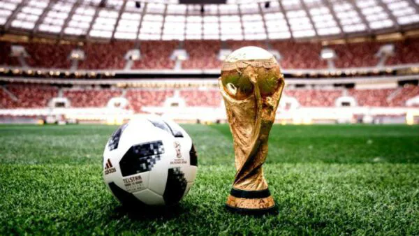 Em primeiro plano, a taça da Copa do Mundo e uma bola de futebol estão sobre o gramado de um estádio. Ao fundo, arquibancadas repletas de torcedores aparecem desfocadas, destacando os dois objetos.