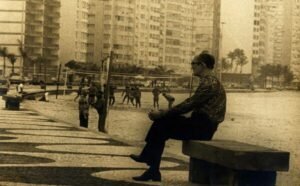 DRUMMOND: FOI-SE A COPA? NÃO FAZ MAL 2 Fotografia em preto e branco de Carlos Drummond de Andrade sentado em um banco à beira da praia de Copacabana. Ao fundo, aparecem pessoas na areia, quadras esportivas, prédios altos e coqueiros.