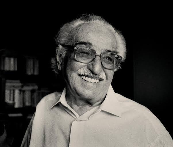 Retrato em preto e branco do poeta Manoel de Barros. Ele sorri, usa óculos de armação grande, camisa clara e bigode. Ao fundo, um ambiente interno desfocado