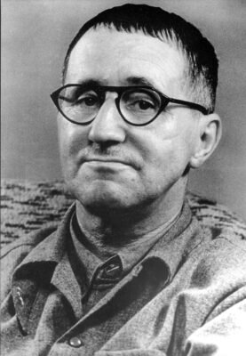 Retrato em preto e branco de Bertolt Brecht. O dramaturgo aparece em primeiro plano, usando óculos de armação escura e camisa de botões. Olha para a câmera com expressão serena e discreto sorriso.