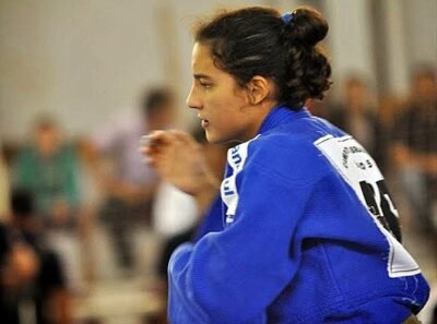 LAtleta de judô aparece de perfil, com os cabelos presos em um coque e vestindo quimono azul. Em posição de concentração, ela observa a competição enquanto o público desfocado aparece ao fundo.