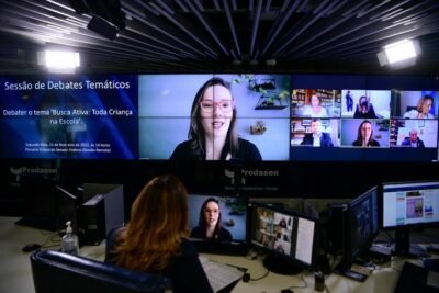 Malala Pacheco participa de uma videoconferência exibida em um grande painel de vídeo durante uma sessão de debates temáticos. Ao lado, outros participantes aparecem em janelas virtuais, enquanto a transmissão é acompanhada de uma sala de controle.
