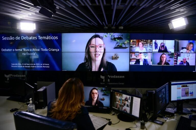 Malala Pacheco participa de uma videoconferência exibida em um grande painel de vídeo durante uma sessão de debates temáticos. Ao lado, outros participantes aparecem em janelas virtuais, enquanto a transmissão é acompanhada de uma sala de controle.