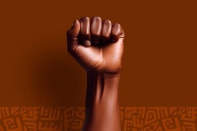 Punho cerrado de uma pessoa negra é erguido diante de um fundo marrom. Na parte inferior, há grafismos em tons mais escuros. A imagem simboliza resistência, luta, força e afirmação da população negra.