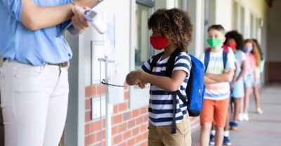 Crianças usam máscaras de proteção e aguardam em fila na entrada de uma escola. Uma profissional mede a temperatura de um estudante com um termômetro infravermelho antes da entrada no prédio.