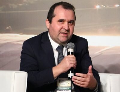 Rubens Valente está sentado durante um evento, com um microfone na mão. Veste terno azul-marinho, camisa branca e gravata cinza. Gesticula enquanto fala e mantém o olhar voltado à frente. Ao fundo, painel desfocado.