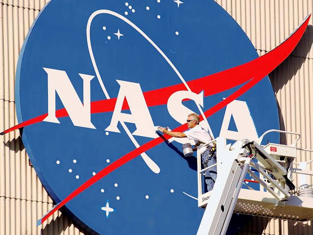 James Webb aparece em uma plataforma elevatória, com um dos braços estendido em direção ao logotipo da NASA. Veste camiseta branca, calça jeans, boné e óculos escuros. Ao fundo, o emblema da agência espacial ocupa quase toda a fachada do edifício.