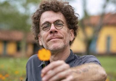 Nando Reis, cantor, aparece em primeiro plano usando óculos redondos e camisa cinza. Com expressão serena, estende a mão em direção à câmera oferecendo uma flor amarela. Ao fundo, jardim e construções desfocadas.