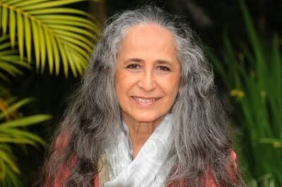 Maria Bethânia aparece em primeiro plano, sorrindo para a câmera. Tem cabelos longos e grisalhos, soltos sobre os ombros. Veste roupa em tom rosado e um lenço branco no pescoço. Ao fundo, folhas e plantas verdes desfocadas compõem o ambiente.