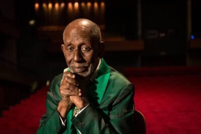 Nelson Sargento aparece sentado, com as mãos entrelaçadas sob o queixo e expressão serena. Veste paletó verde e camisa branca. Ao fundo, um teatro com carpete vermelho e iluminação suave cria um ambiente intimista.
