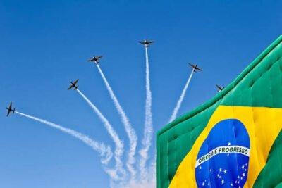 Cinco aeronaves sobrevoam o céu azul em formação, deixando rastros brancos de fumaça que se abrem em leque. À direita, parte da bandeira do Brasil aparece em destaque, com as cores verde, amarela e azul visíveis.