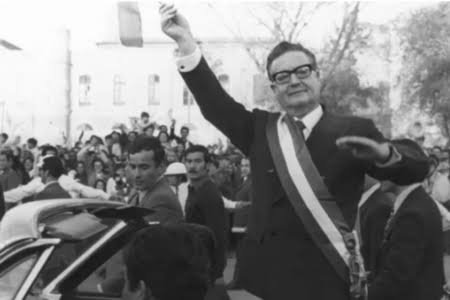 "Homem aparece em pé em um carro conversível, com um dos braços erguidos em gesto de saudação à multidão. Veste terno escuro, faixa presidencial e óculos. Ao fundo, dezenas de pessoas acompanham o desfile em uma rua arborizada. A imagem está em preto e branco.