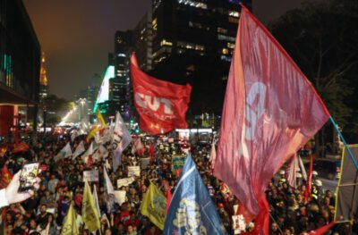 Manifestação ocupa uma avenida à noite. Centenas de pessoas carregam bandeiras de sindicatos e movimentos sociais. Em destaque, bandeiras vermelhas da CUT são erguidas entre os participantes, com prédios iluminados ao fundo.