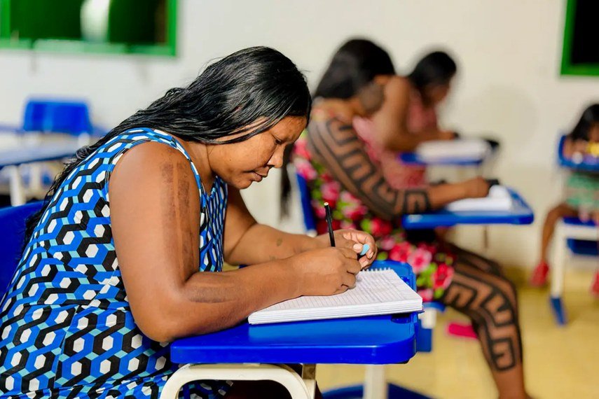 Mulher indígena sentada em carteira escolar azul escreve em um caderno. Ao fundo, outras mulheres também estudam em sala de aula. Uma delas exibe pinturas corporais geométricas nos braços e pernas.