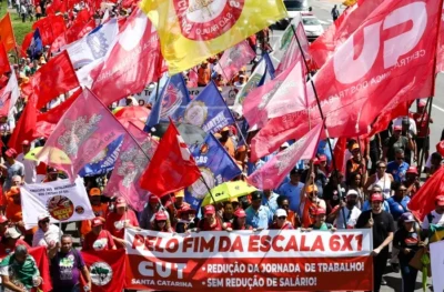 FIM DA ESCALA 6X1: PRESSIONE OS SENADORES E SENADORAS 1 Multidão participa de manifestação em via pública, carregando bandeiras de sindicatos e movimentos sociais. Em destaque, uma faixa vermelha traz a mensagem: “Pelo fim da escala 6x1”. Ao fundo, centenas de pessoas seguem reunidas em defesa da redução da jornada de trabalho sem redução salarial.