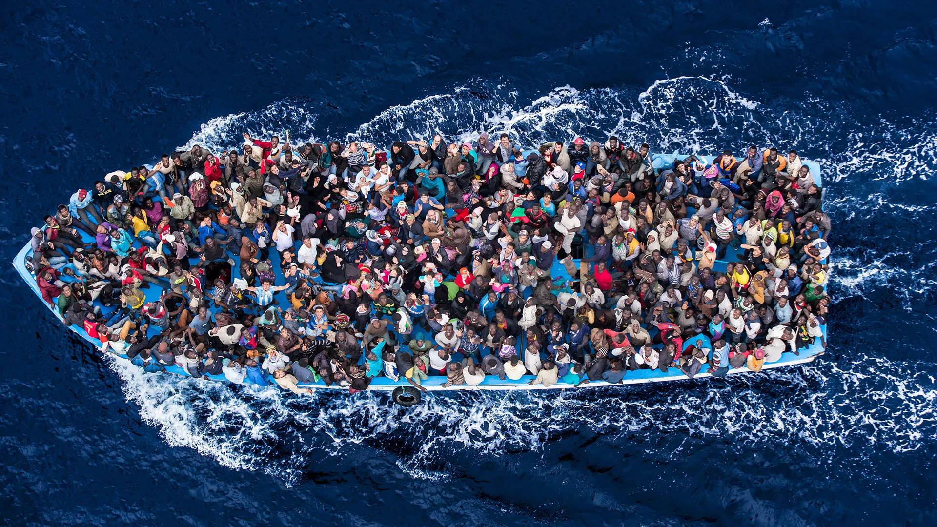 Vista aérea de uma embarcação superlotada em alto-mar, com centenas de pessoas espremidas a bordo. Ao redor, o oceano azul evidencia a precariedade da travessia e os riscos enfrentados por migrantes e refugiados.