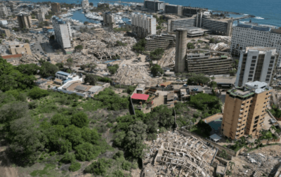 VENEZUELA ENTRE ESCOMBROS: TERREMOTOS AGRAVAM CRISE 1 Vista aérea de uma área urbana com diversos edifícios parcialmente destruídos e grandes montes de escombros. Ao redor, há prédios preservados, áreas com vegetação e o mar visível ao fundo.
