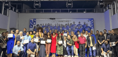 oto de grupo em um palco com dezenas de pessoas reunidas após uma cerimônia. Algumas seguram certificados. Ao fundo, há um painel com a identidade visual do Sindicato dos Bancários de Brasília.