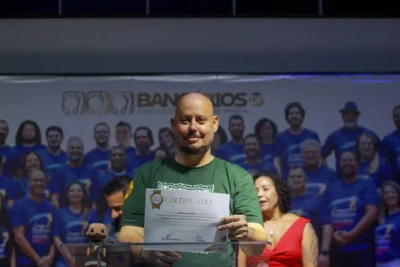 Rodrigo Brito, presidente do Sindicato dos Bancários de Brasília, está em pé, sorrindo e segurando um certificado. Ao fundo, há um palco com pessoas reunidas e um painel com a identidade visual do sindicato.