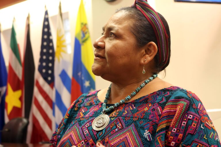 Rigoberta Menchú aparece em primeiro plano, de perfil, com o olhar voltado para cima e expressão serena. Veste roupa com grafismos coloridos, colar de contas turquesa com pingente e faixa multicolorida nos cabelos. Ao fundo, bandeiras de diferentes países aparecem desfocadas.