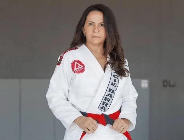 HOME 4 Mulher aparece em pé, com expressão séria e olhar voltado à frente. Veste quimono branco de jiu-jítsu, faixa vermelha e preta na cintura e uma faixa com a inscrição "Gracie Barra". Mantém as mãos apoiadas sobre a faixa. Ao fundo, um ambiente interno com paredes em tons de cinza aparece desfocado. O CORPO FEMININO E A POTÊNCIA NEGADA